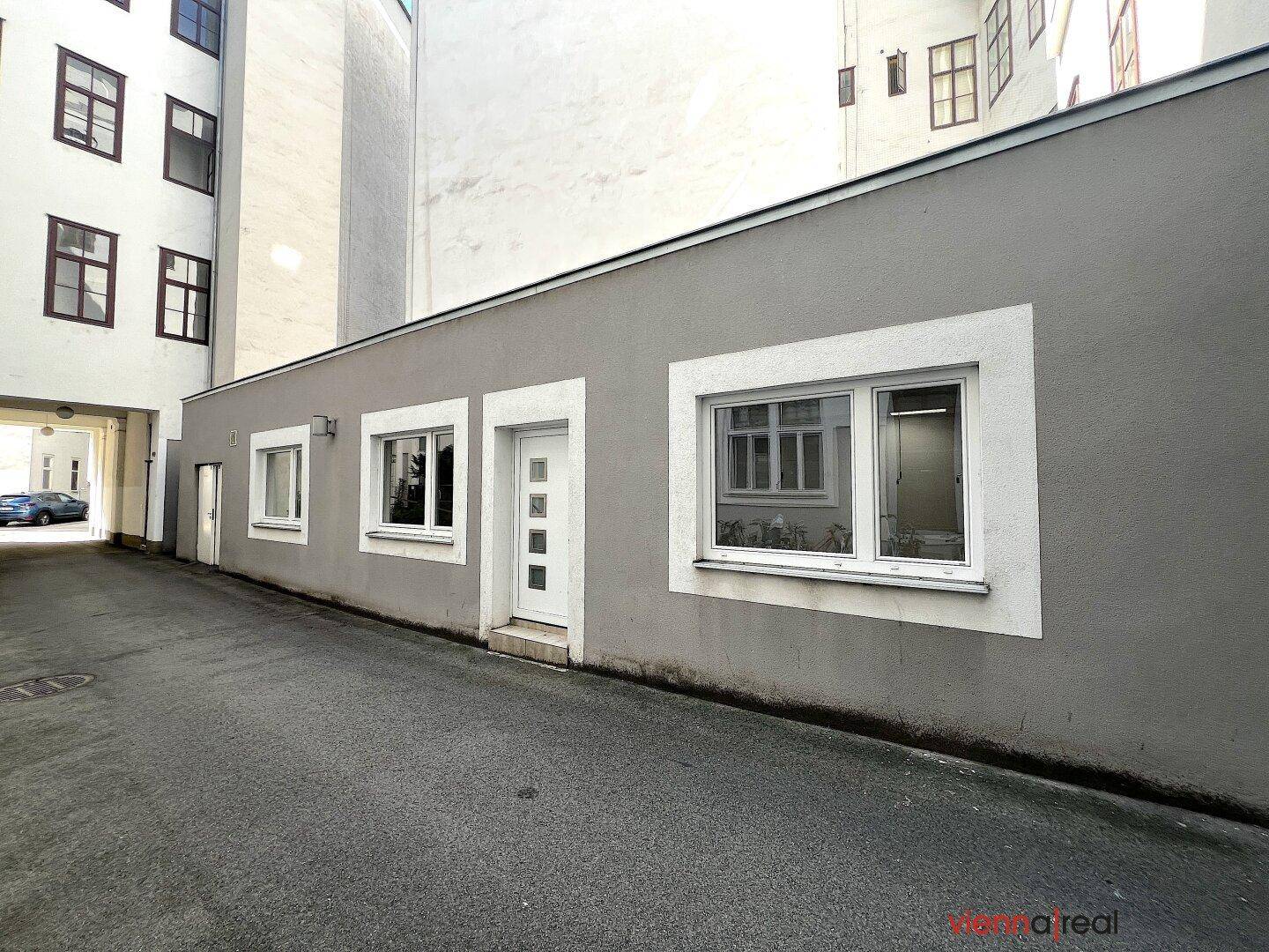 Graue Gebäudefassade mit weißen Fenstern und Tür im Innenhof, umgeben von weiteren Gebäuden.