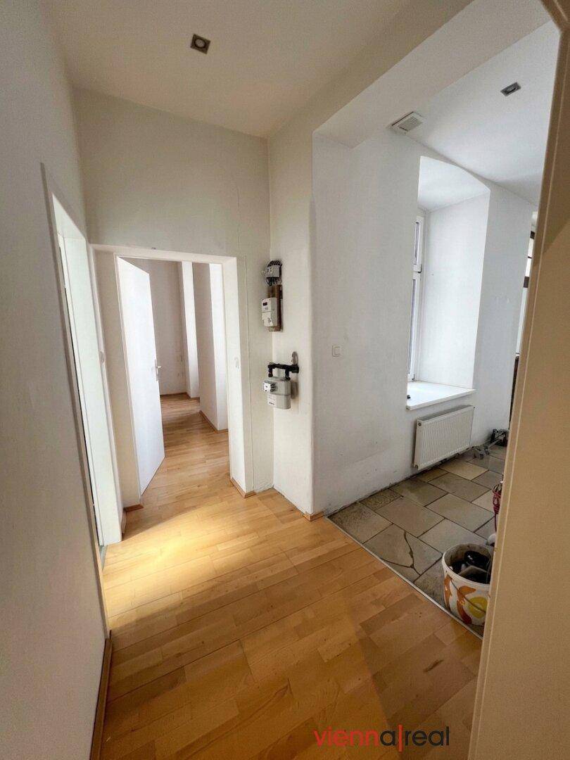 Flur mit Holzboden, der in einen gefliesten Bereich mit Fenster und Heizkörper übergeht.