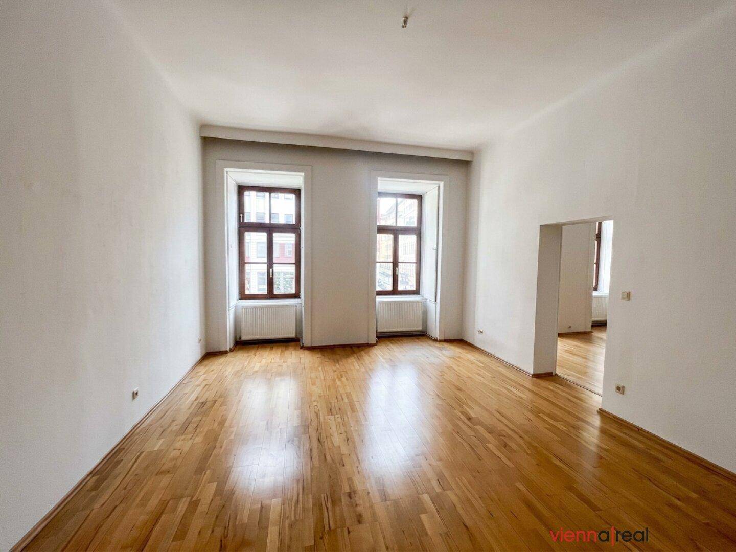 Leeres Zimmer mit Parkettboden und zwei Fenstern, die viel Tageslicht hereinlassen.