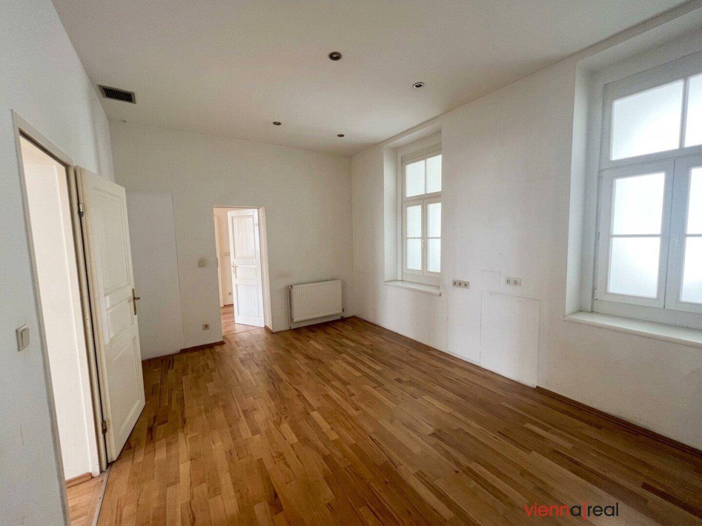 Helles Zimmer mit Parkettboden und zwei Fenstern, das einen angenehmen Wohnraum bietet.