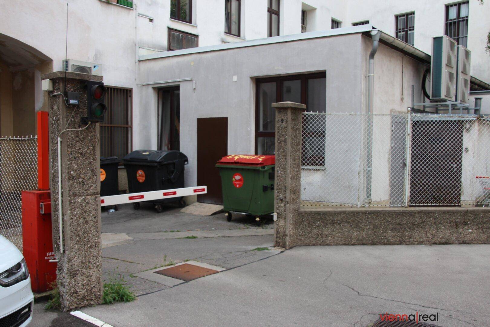 Eingangsbereich zum Innenhof mit Schranke, Müllcontainern und einem kleinen Nebengebäude.