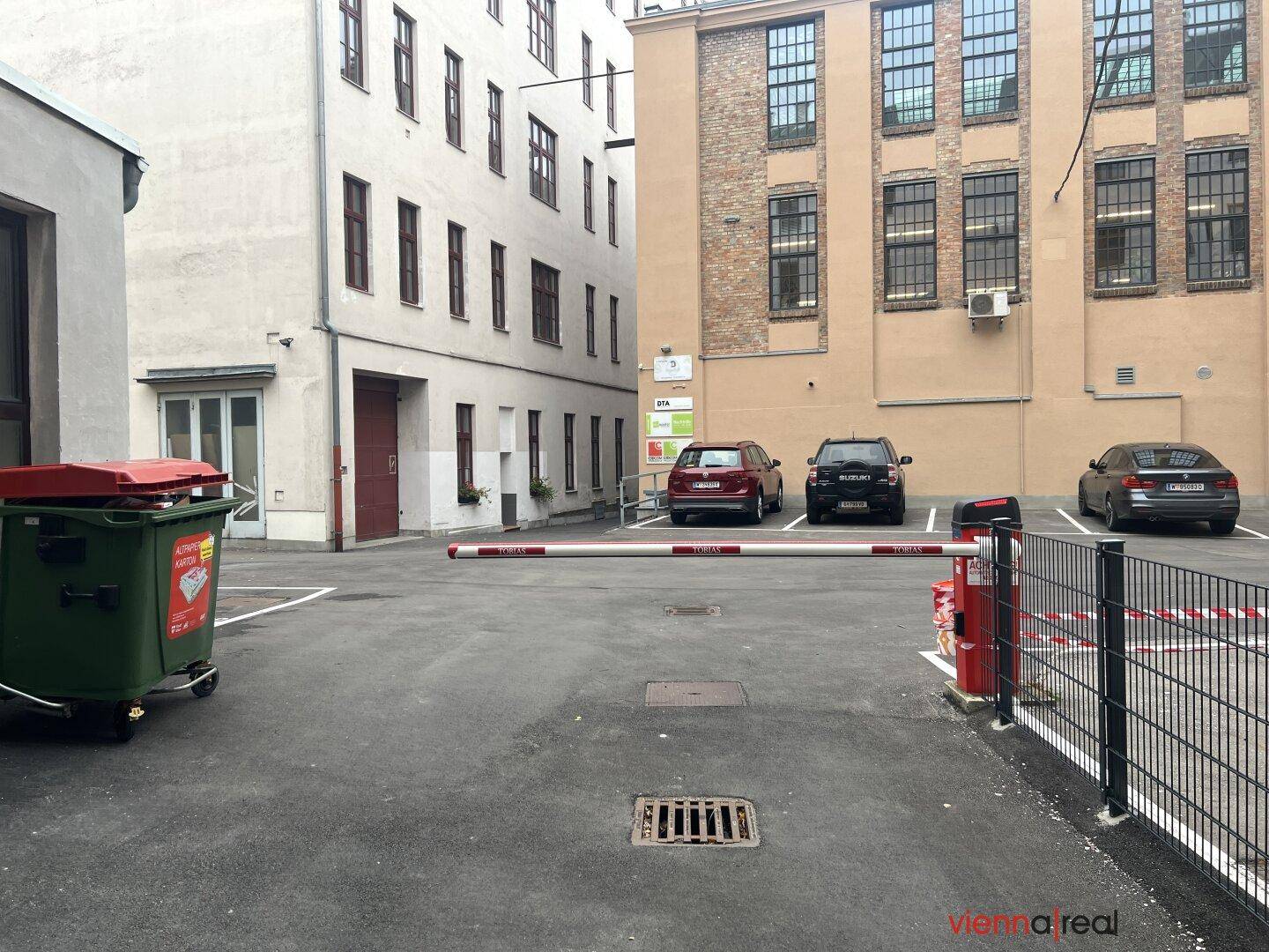 Zufahrt zum Innenhofparkplatz mit Schranke, Mülltonnen und umliegenden Gebäuden.