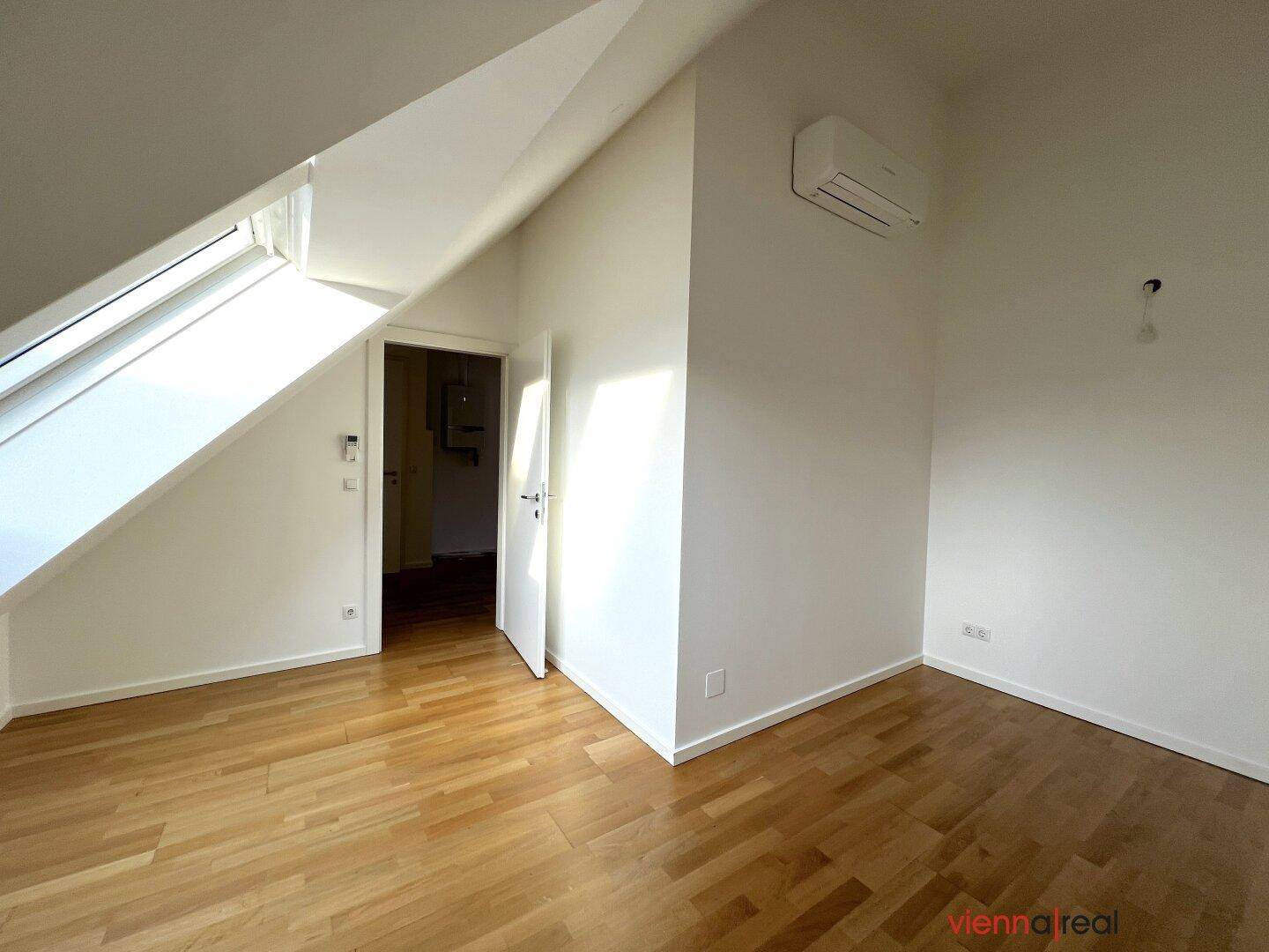 Heller Raum mit Holzboden, Klimaanlage und offener Tür, unter einem schrägen Dach.