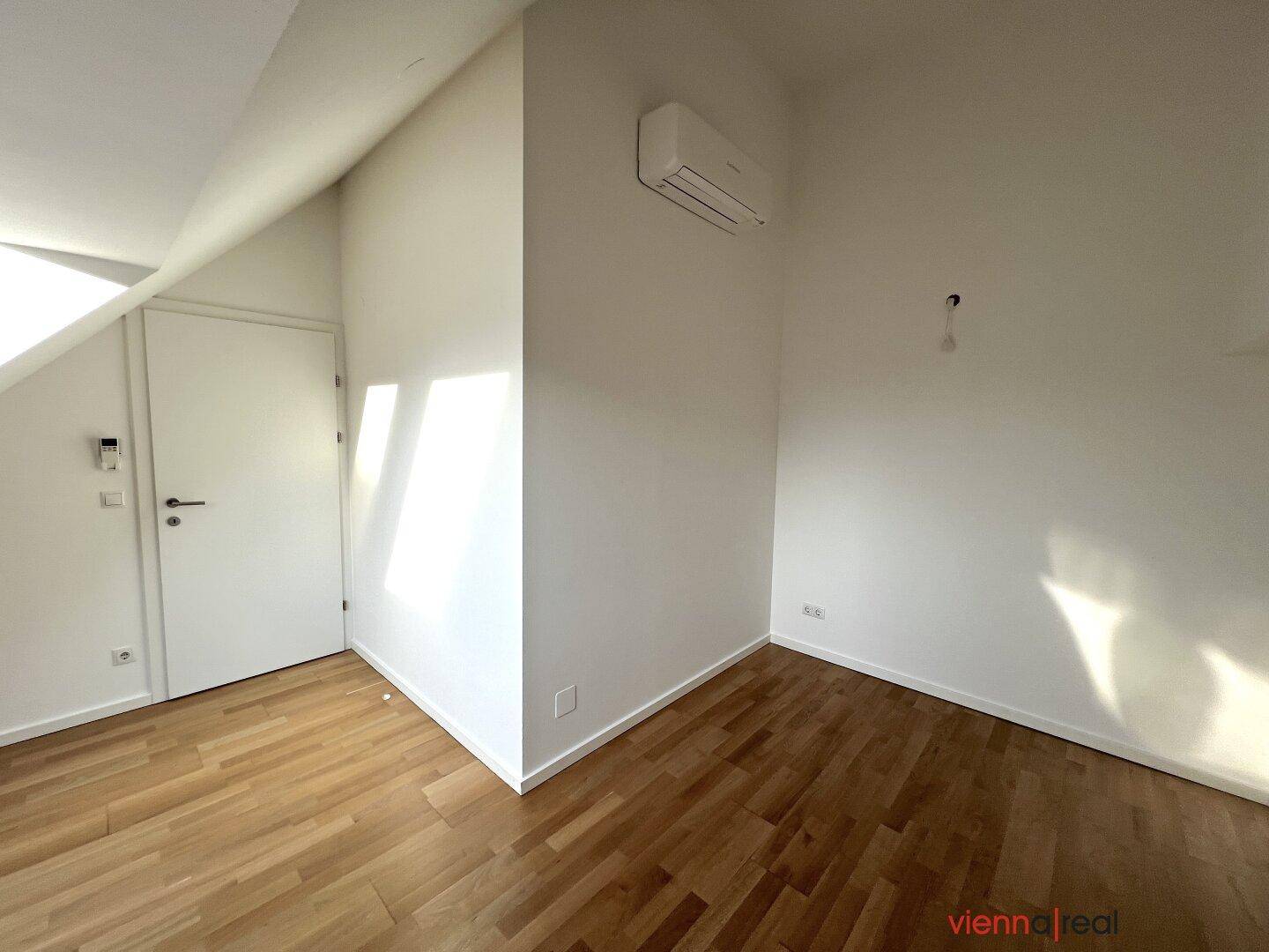 Leerer Raum mit Holzboden, Klimaanlage und geschlossener Tür, unter einem schrägen Dach.