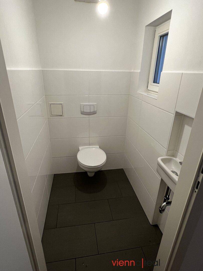 Gäste-WC mit weißen Fliesen, Toilette, kleinem Waschbecken und einem Fenster.
