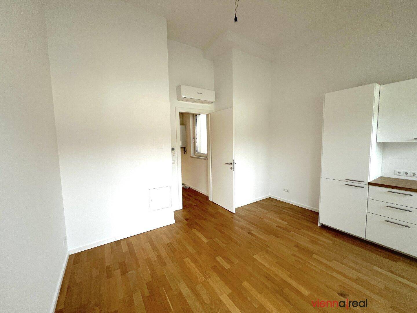 Leerer Raum mit Holzboden, Klimaanlage und offener Tür, mit Blick auf die angrenzende Küche.