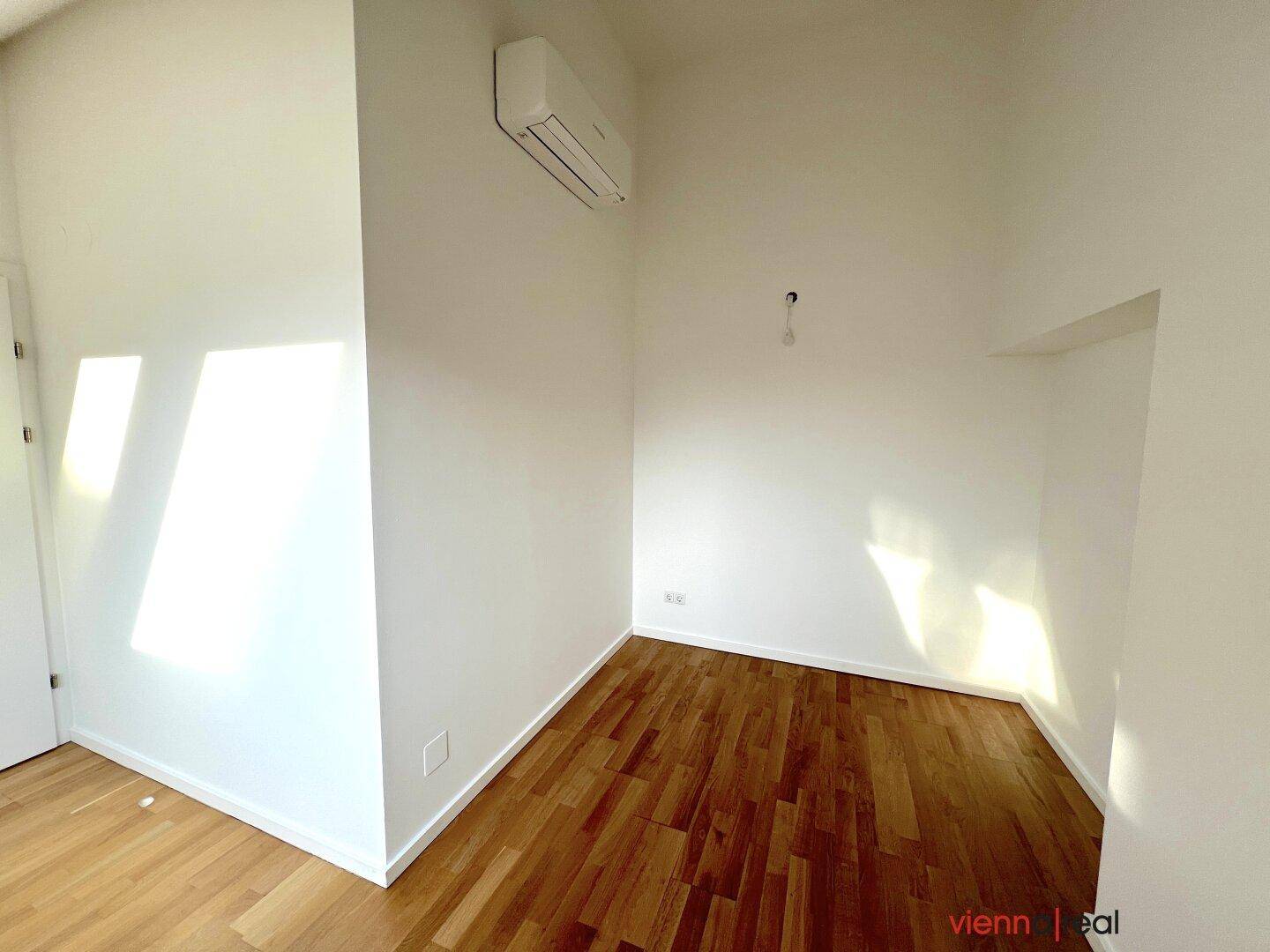 Heller Raum mit Holzboden, Klimaanlage und einer Nische in der Wand, unter einem schrägen Dach.