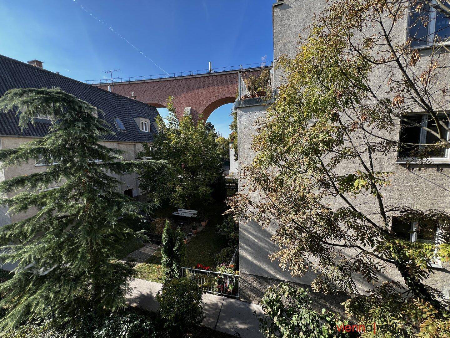 Blick auf einen begrünten Innenhof und eine historische Brücke im Hintergrund an einem sonnigen Tag.