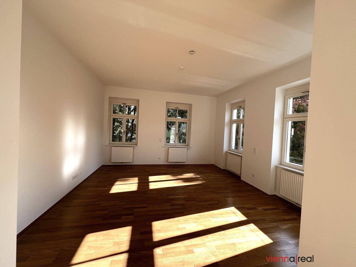 Helles Zimmer mit Parkettboden und mehreren Fenstern, die viel Tageslicht hereinlassen.