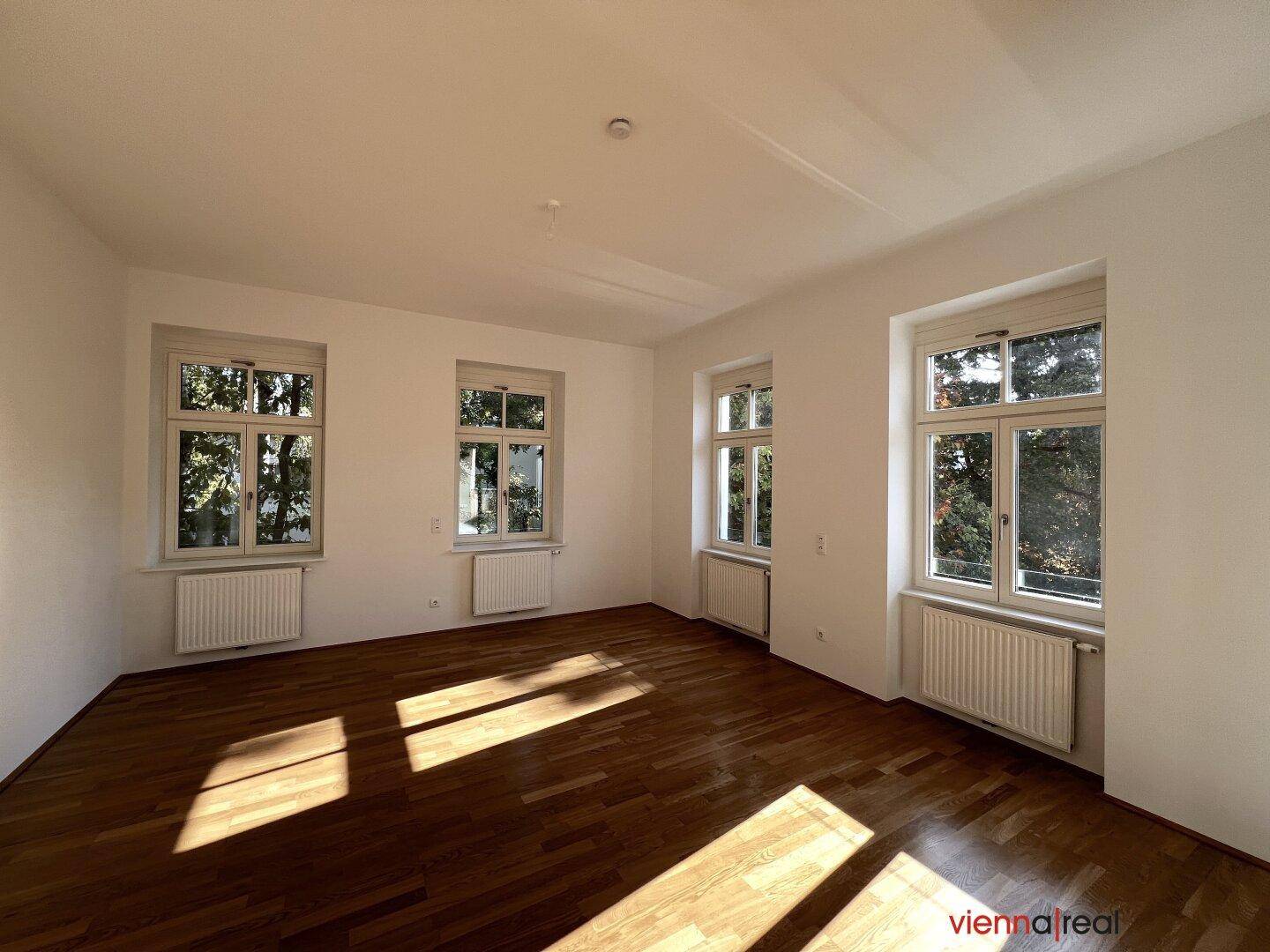 Lichtdurchflutetes Zimmer mit Parkettboden und Blick ins Grüne durch die großen Fenster.