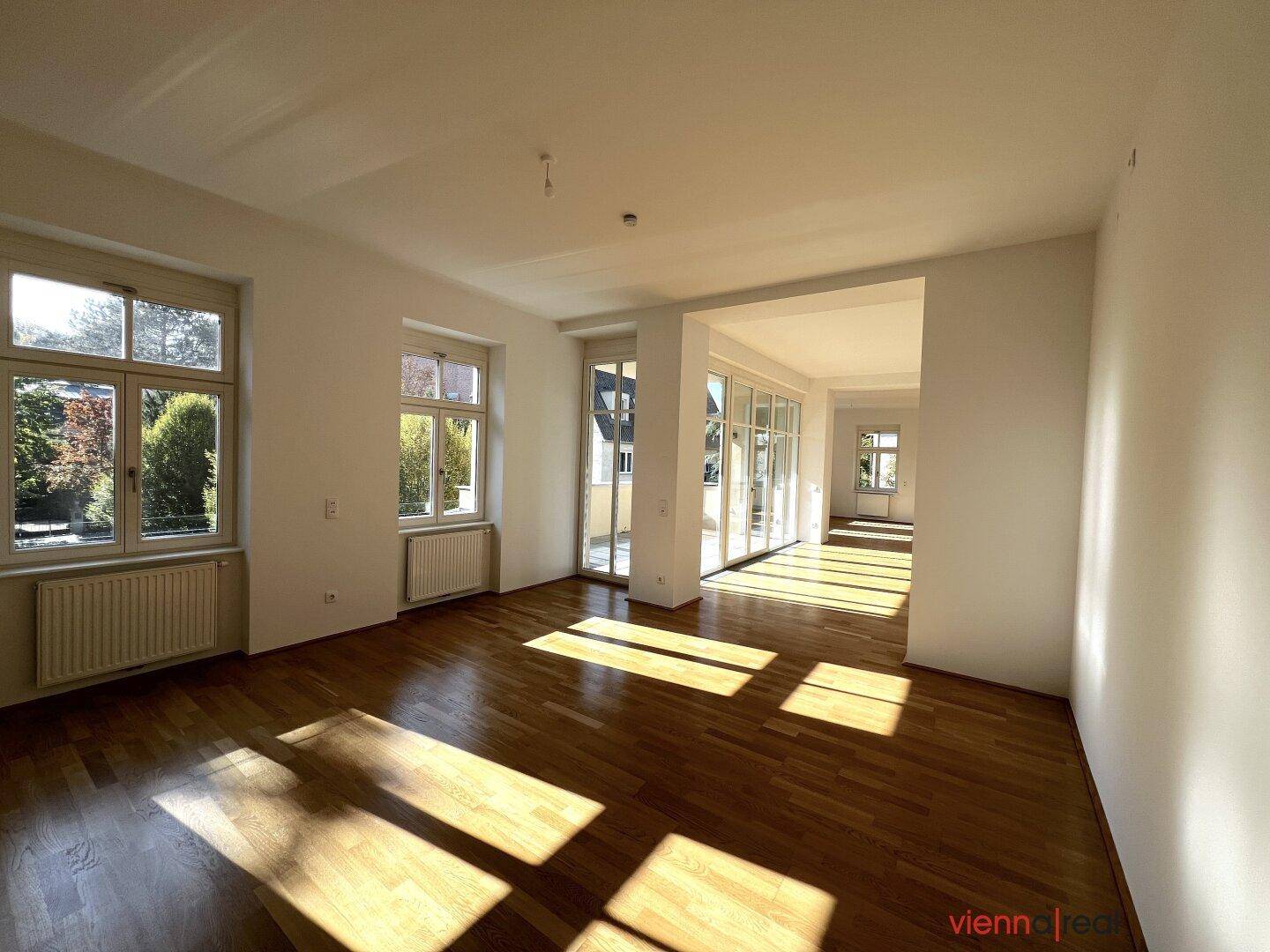 Geräumiger Wohnbereich mit Parkettboden, großen Fenstern und Zugang zu einem Balkon.