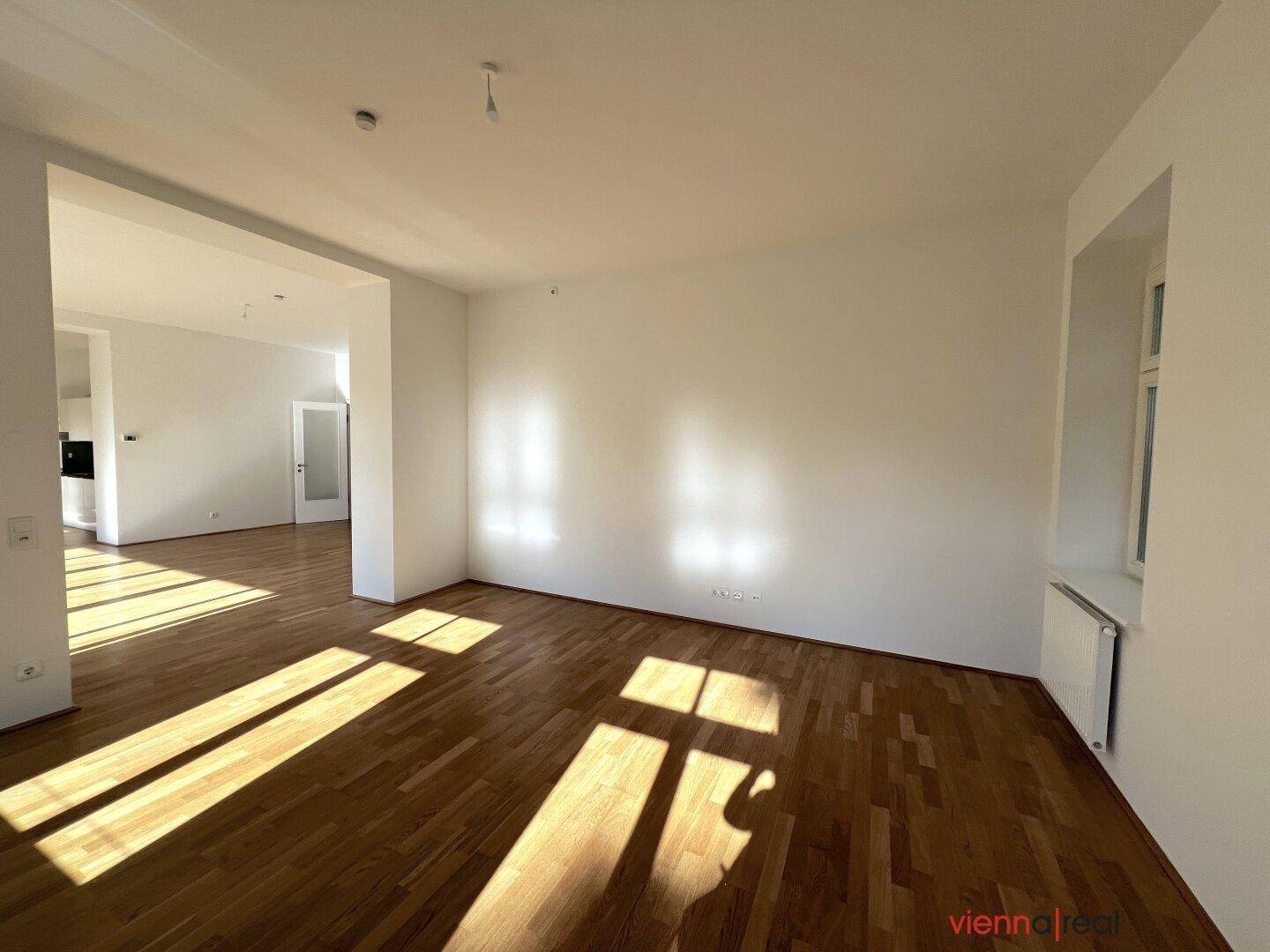 Heller, leerer Raum mit Parkettboden und sonnigen Lichtflecken auf dem Boden.