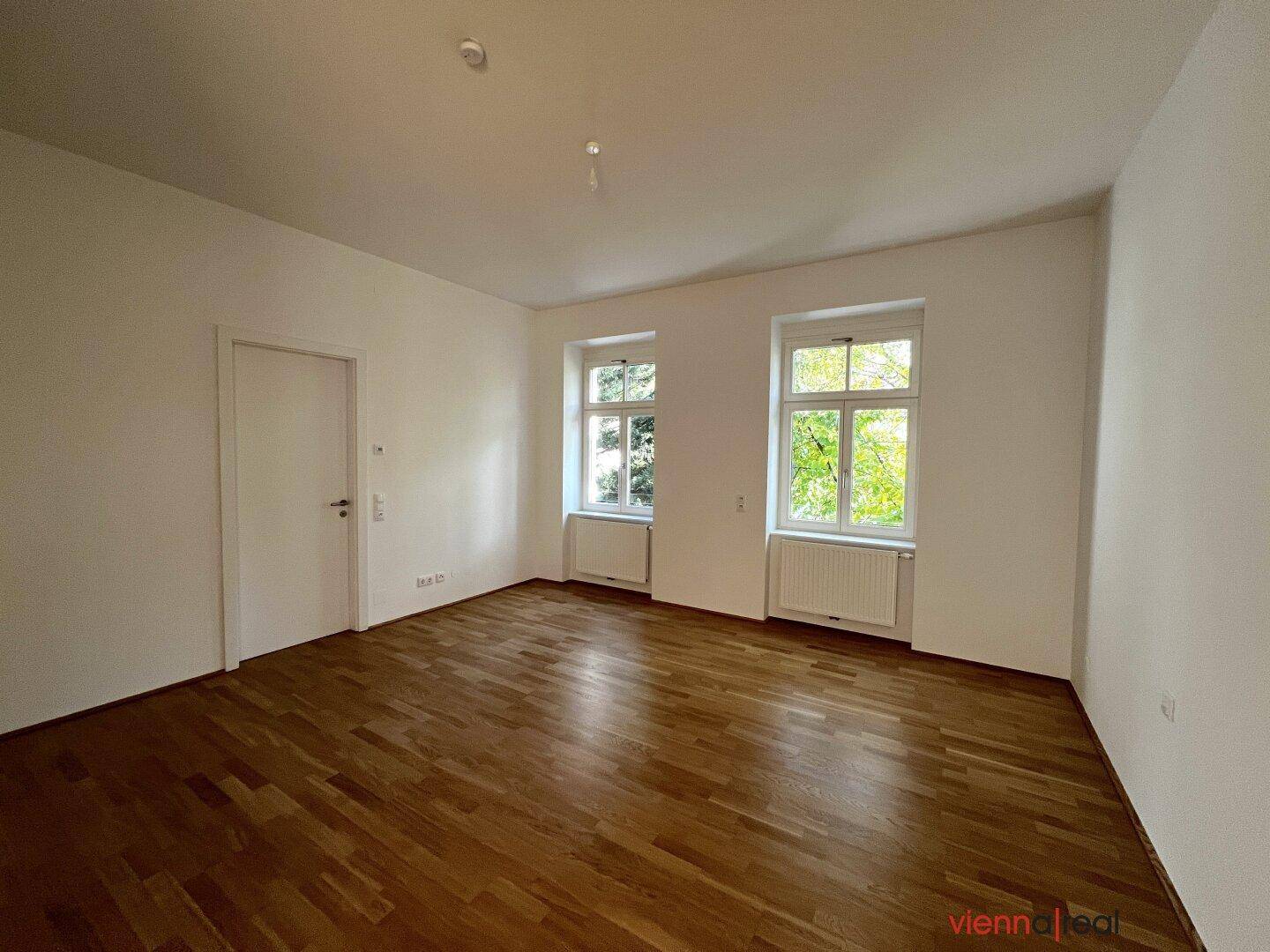 Helles Zimmer mit Parkettboden und zwei Fenstern, die einen Blick ins Grüne bieten.