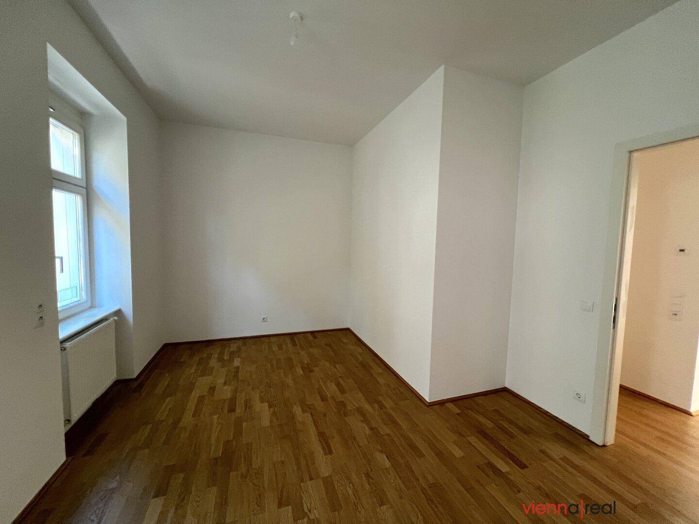 Geräumiges Zimmer mit Parkettboden, einem Fenster und Zugang zu einem weiteren Raum.