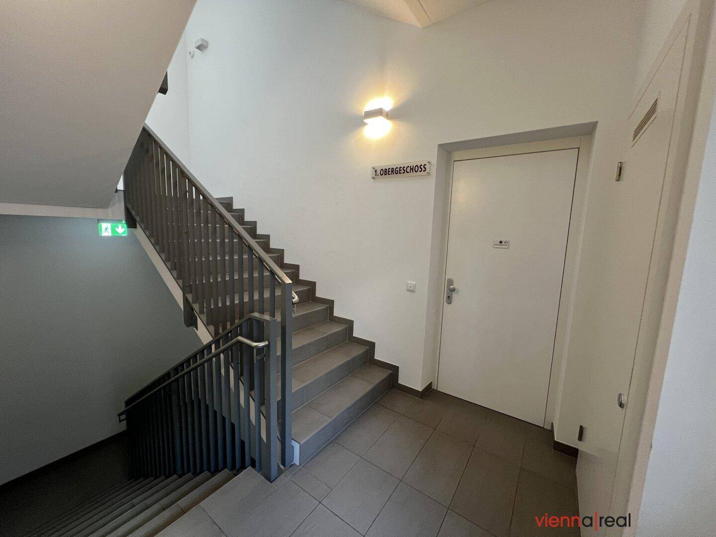 Modernes Treppenhaus mit Geländer und Beschilderung zum ersten Obergeschoss.
