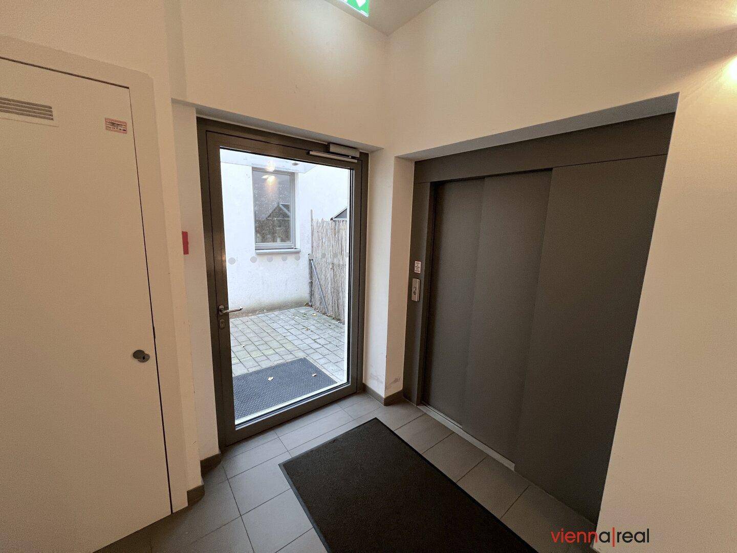 Heller Eingangsbereich mit Fliesenboden, Zugang zum Aufzug und einer Glastür zum Innenhof.