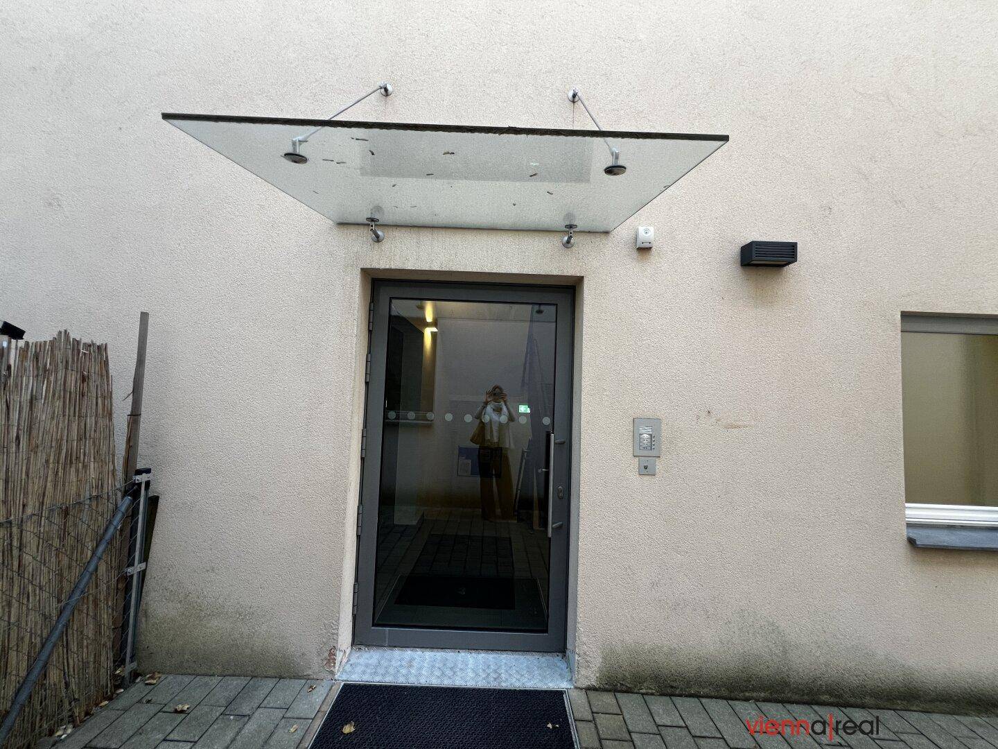 Haupteingang des Gebäudes mit Glasvordach, Gegensprechanlage und gepflastertem Zugangsbereich.