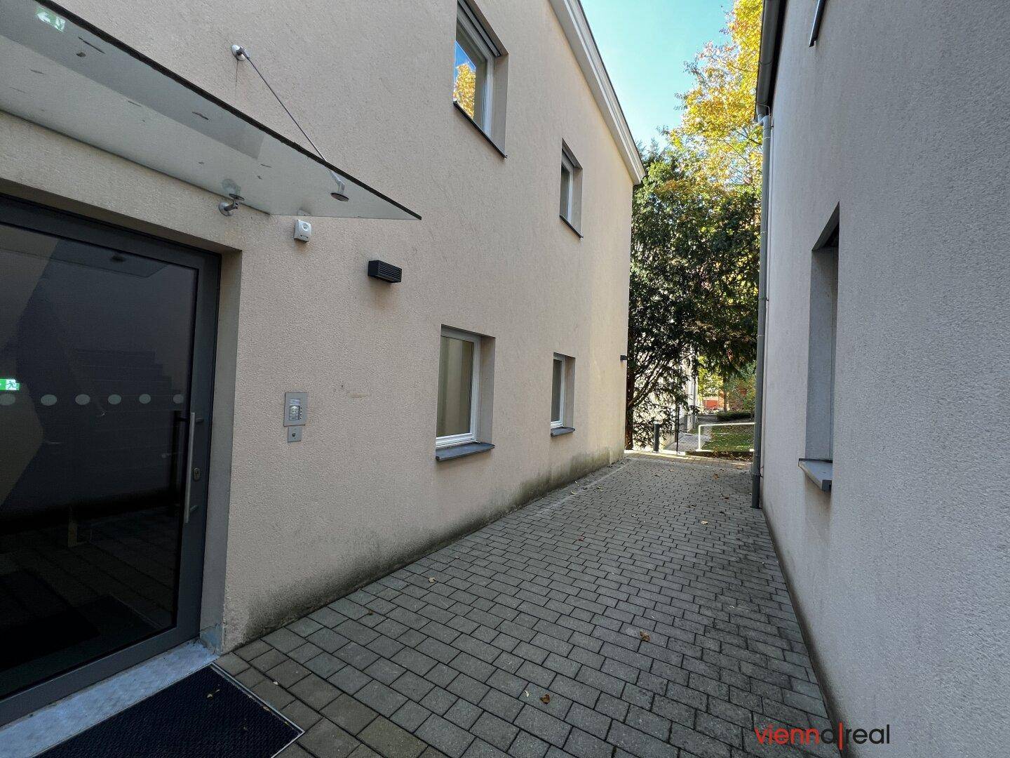 Gepflasterter Weg entlang der Gebäudeseite mit Bäumen und Sträuchern im Hintergrund.