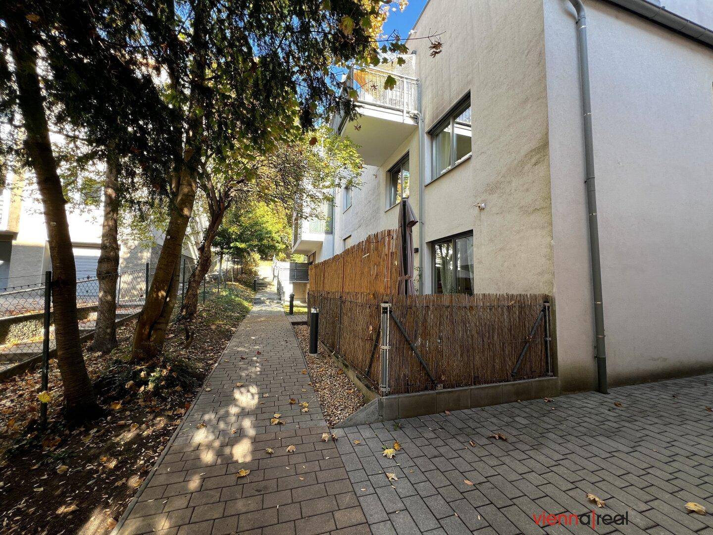 Gepflasterter Gehweg neben dem Gebäude mit einer kleinen, eingezäunten Fläche und Bäumen.