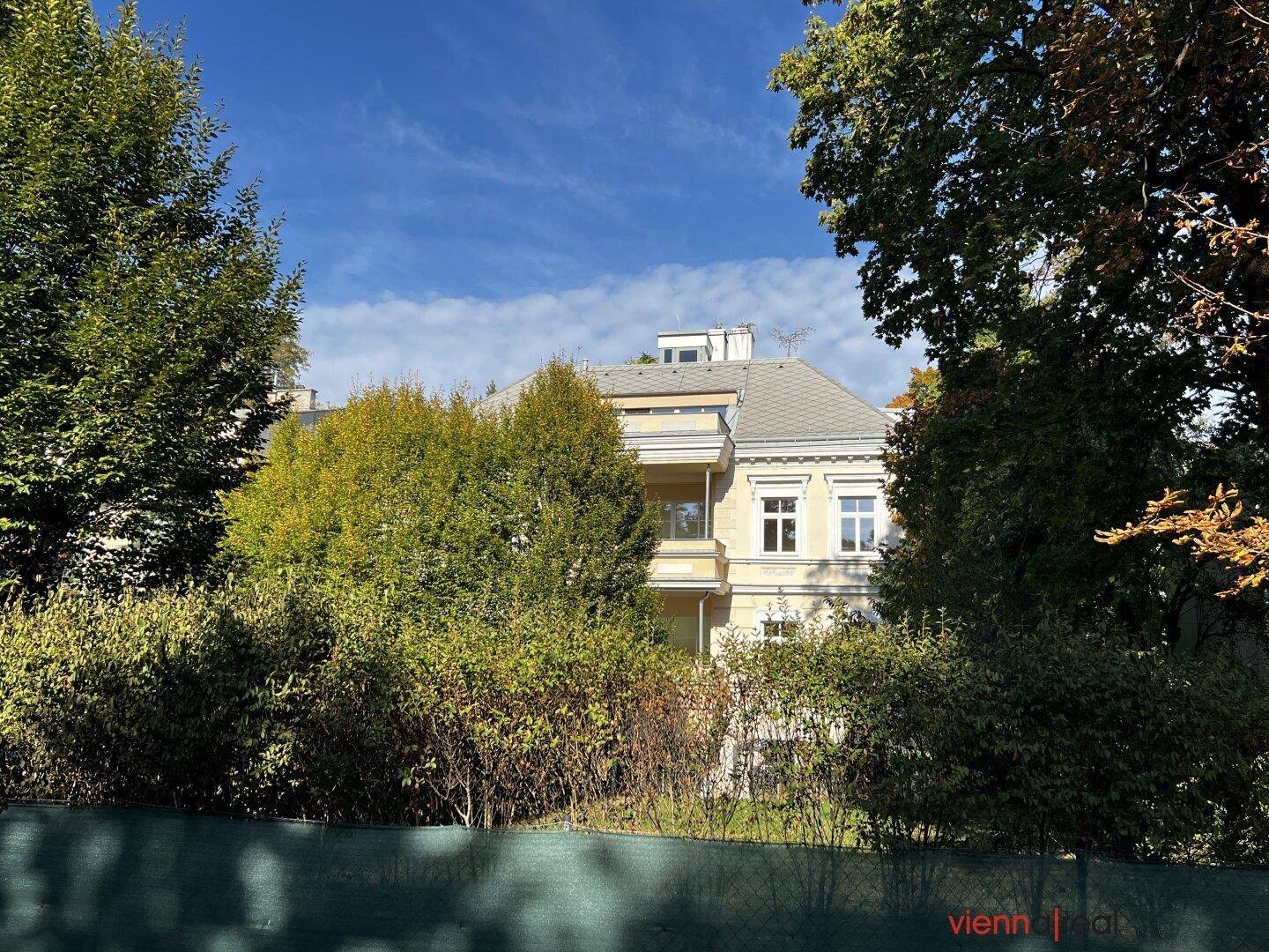 Ansicht des Gebäudes hinter Bäumen und Sträuchern, mit blauem Himmel im Hintergrund.