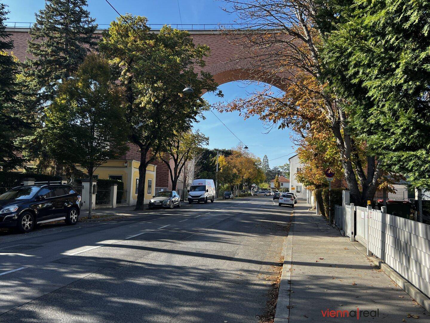 Ruhige Wohnstraße mit Bäumen und einer historischen Brücke im Hintergrund an einem sonnigen Tag.