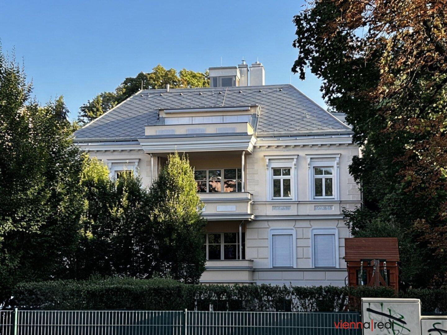 Ansicht der Gebäudefassade mit gepflegter Hecke und Zaun im Vordergrund, umgeben von Bäumen.