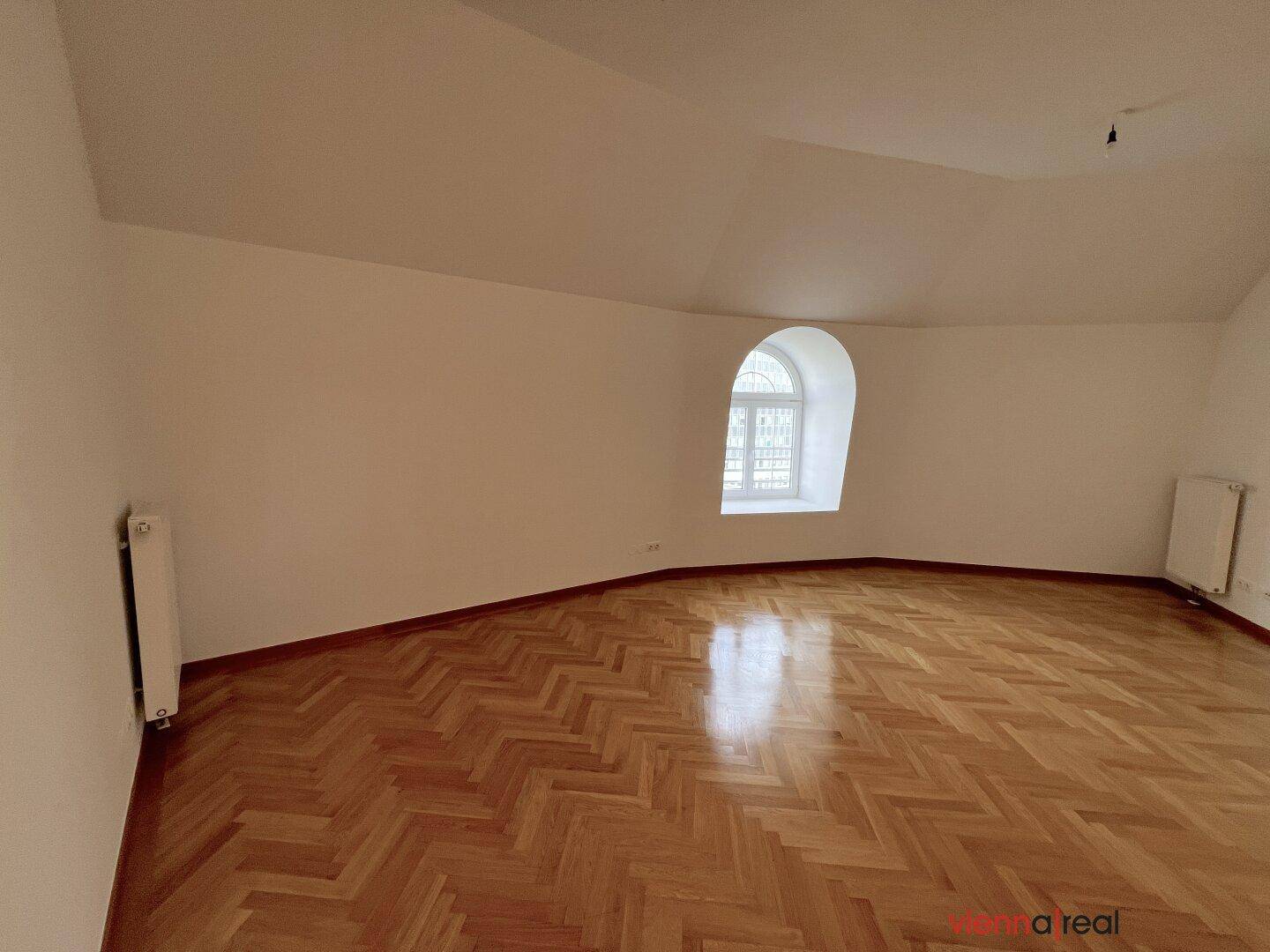 Heller Raum mit Fischgrätparkett, weißen Wänden und einem charmanten Rundbogenfenster.