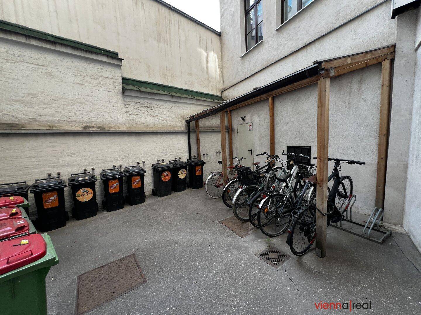 Hinterhof mit Fahrradabstellplatz und Mülltonnen, umgeben von Gebäudewänden.