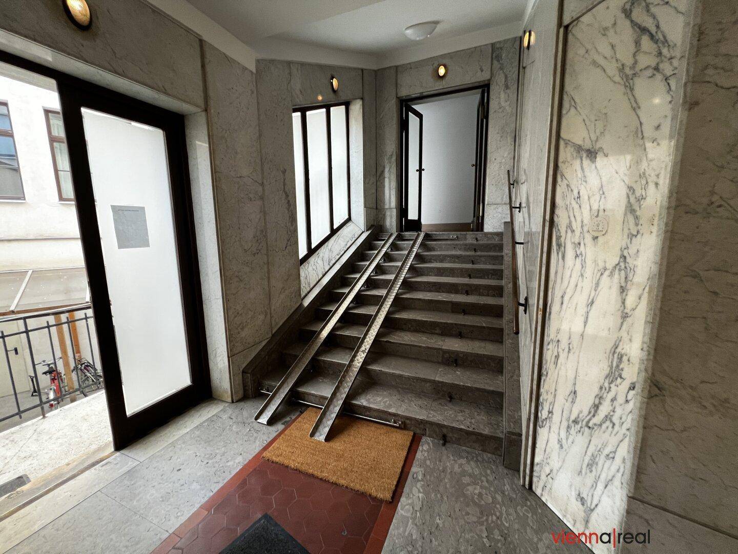 Eingangsbereich mit Marmorverkleidung, Treppe und einer Rollstuhlrampe für Barrierefreiheit.