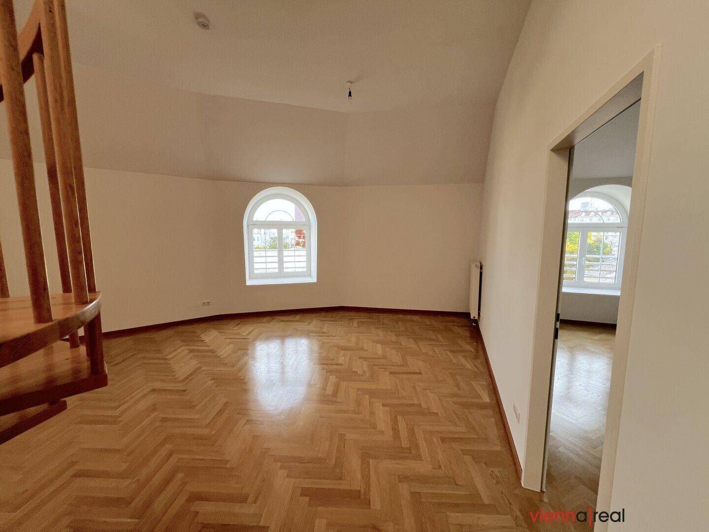 Geräumiger Wohnbereich mit glänzendem Fischgrätparkett, Rundbogenfenster und Zugang zu weiteren Räumen.