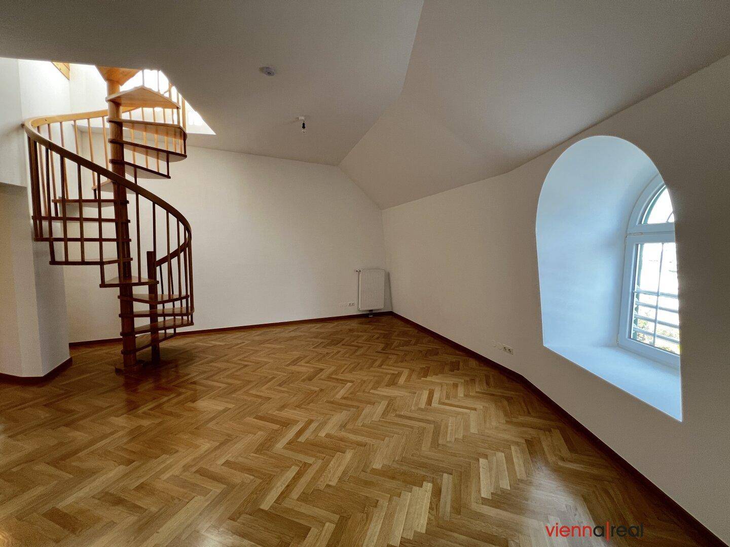 Lichtdurchfluteter Raum mit Fischgrätparkett, Rundbogenfenster und einer dekorativen Holzwendeltreppe.