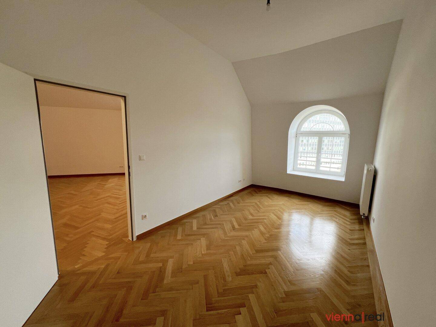 Ruhiger Raum mit glänzendem Fischgrätparkett, weißen Wänden und einem eleganten Rundbogenfenster.