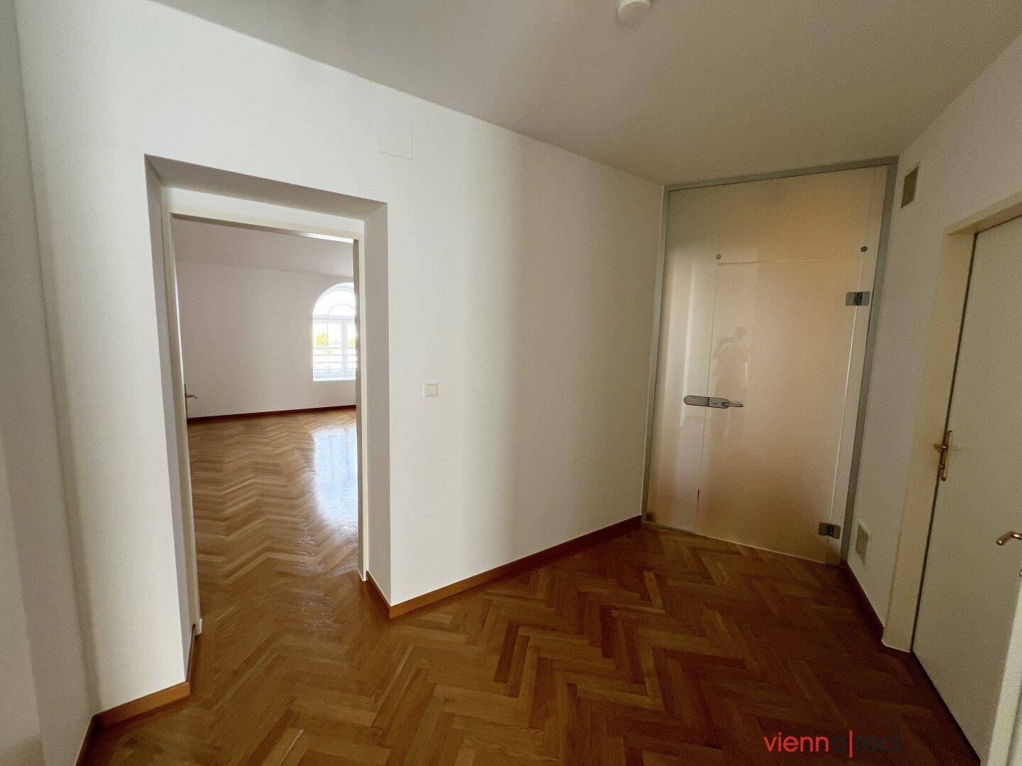Heller Flur mit Fischgrätparkett, Milchglastür und offenem Zugang zu einem weiteren Wohnraum.