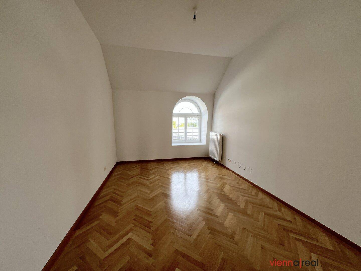 Geräumiges Schlafzimmer mit glänzendem Fischgrätparkett, weißen Wänden und einem Rundbogenfenster.