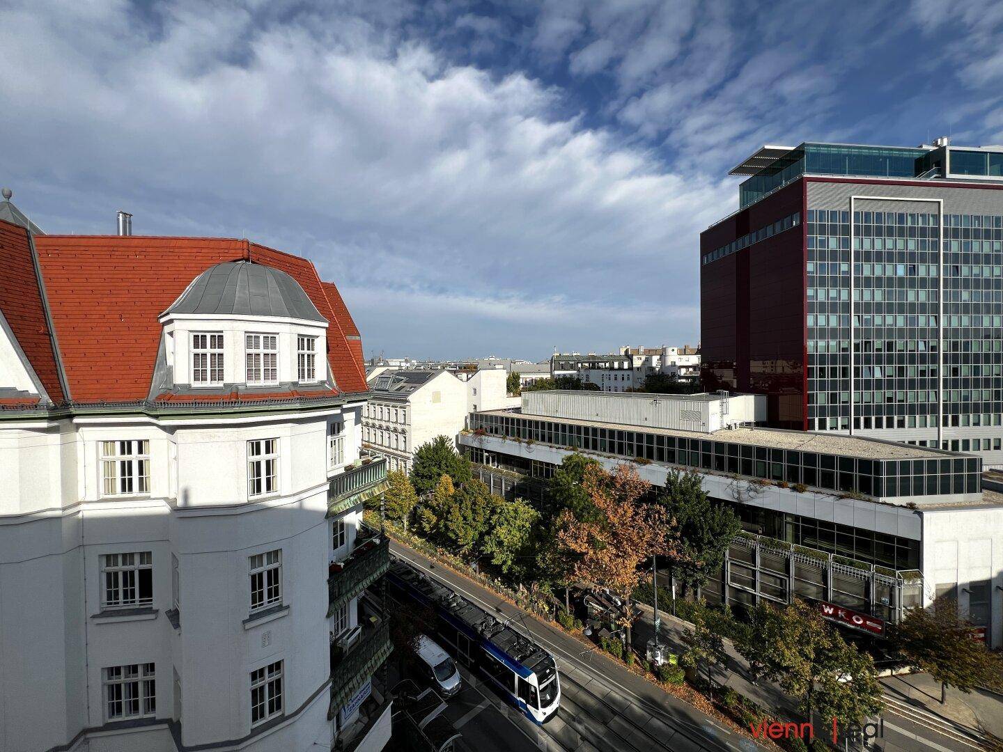 Stadtansicht mit traditionellen und modernen Gebäuden, Bäumen und einer Straßenbahn unter blauem Himmel.