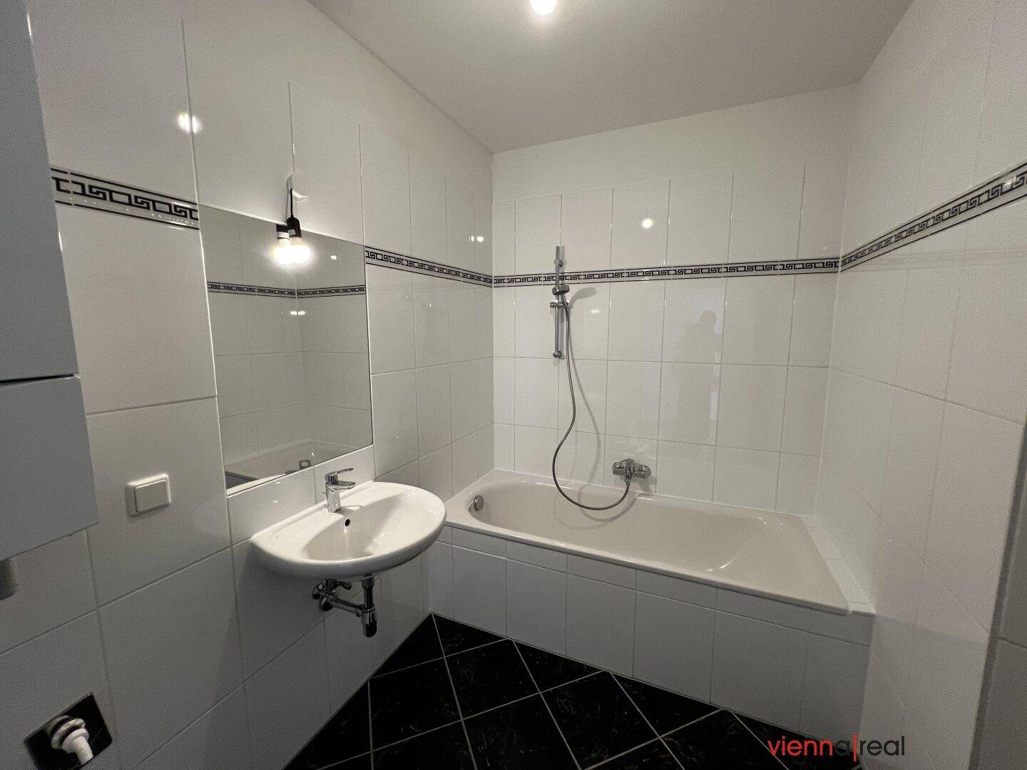 Helles Badezimmer mit weißen Wandfliesen, schwarzen Bodenfliesen, Badewanne und Waschbecken.