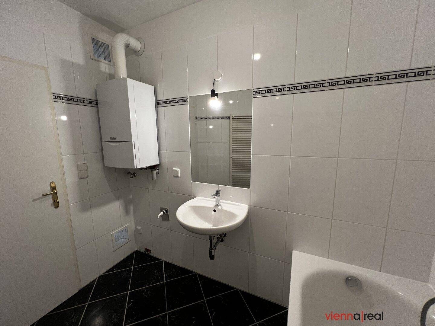Badezimmer mit weißen Wandfliesen, schwarzen Bodenfliesen, Waschbecken und Wandheizkessel.