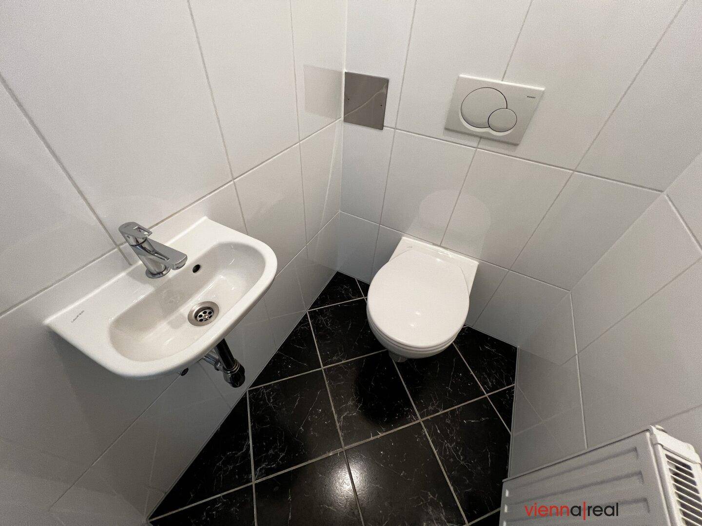 Detailansicht des WCs mit wandhängender Toilette, kleinem Waschbecken und Heizkörper.