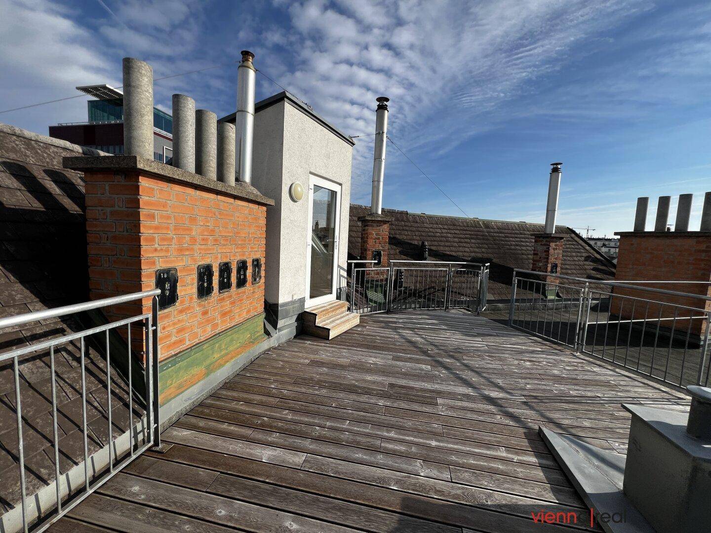 Weitläufige Dachterrasse mit Holzdielen und Blick auf die umliegenden Dächer und den Himmel.