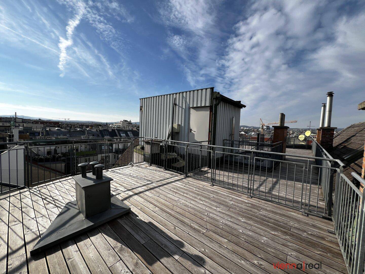 Große Dachterrasse mit Holzdielen und Blick auf ein Nebengebäude und die Stadt.