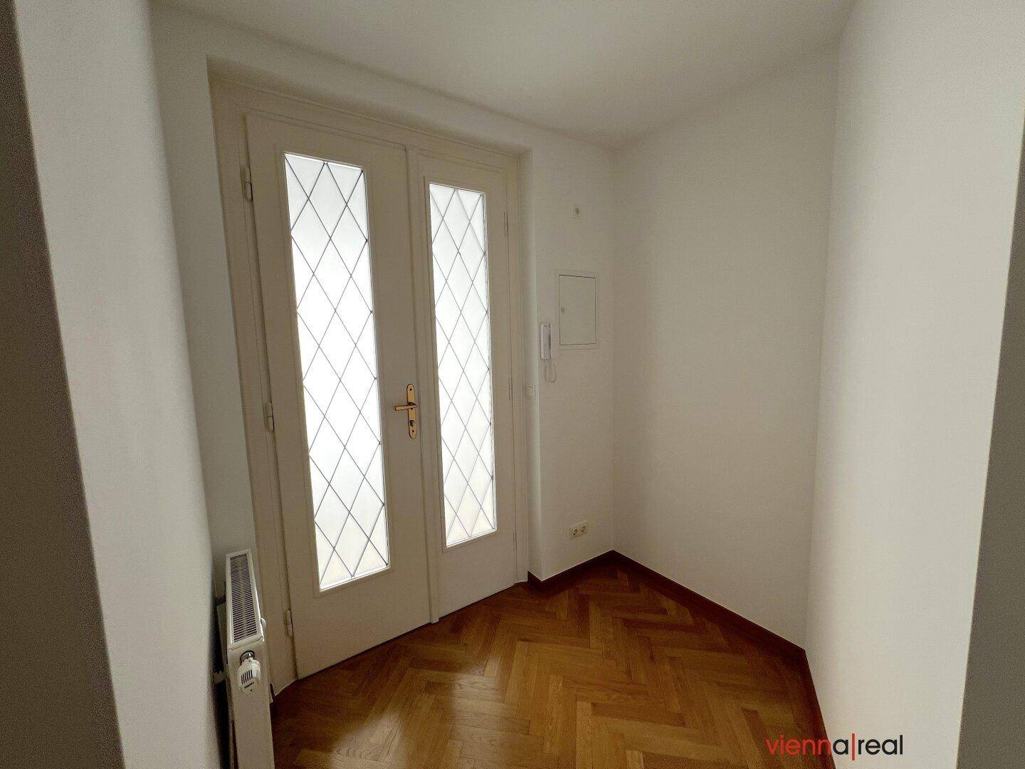 Eingangsbereich mit Fischgrätparkett, Heizkörper und einer Doppeltür mit Glasausschnitten.