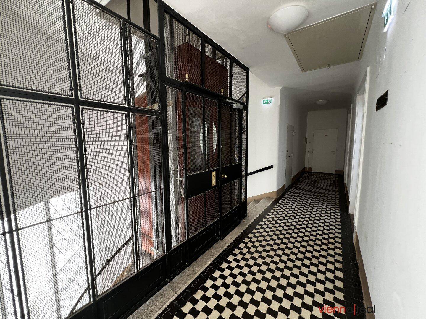 Langer Flur in einem Altbau mit Schachbrettmuster-Fliesenboden und einem historischen Aufzug.