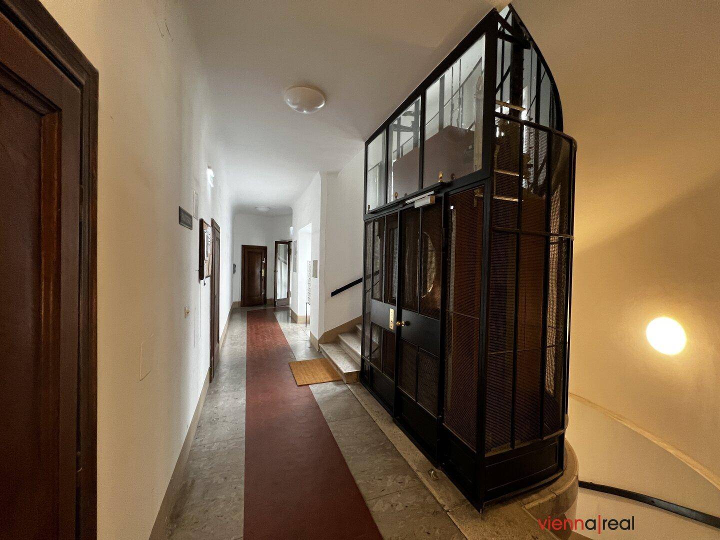 Geräumiger Flur in einem Altbau mit Aufzug, Treppe und Teppichläufer.
