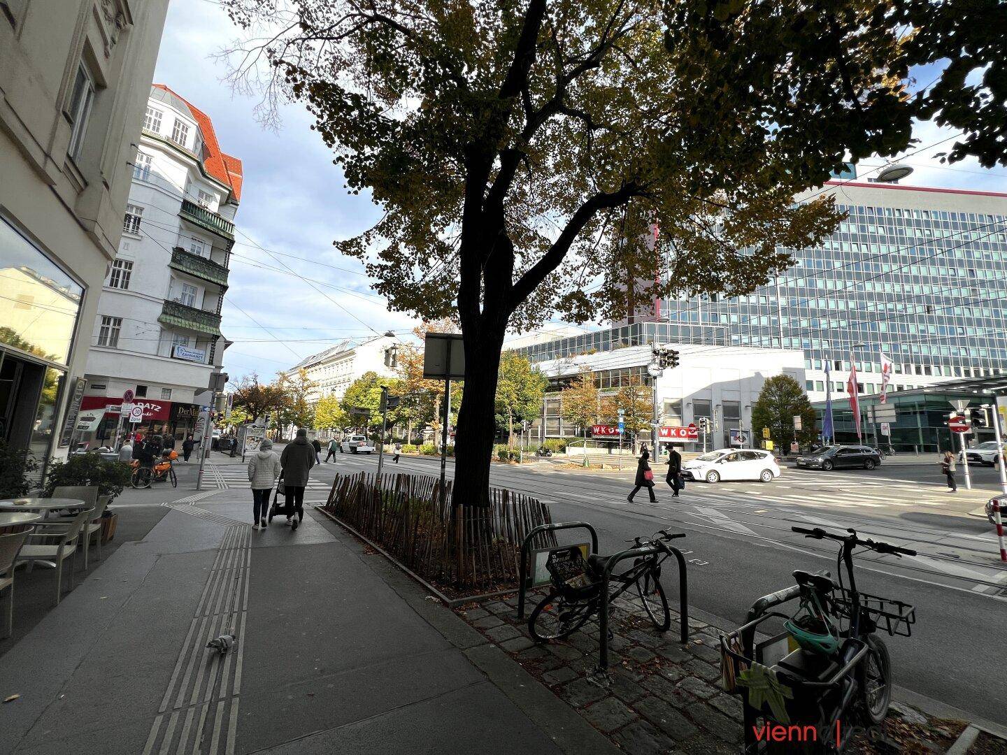 Belebtes Stadtbild mit Fußgängern, Gebäuden und Bäumen an einem sonnigen Tag.