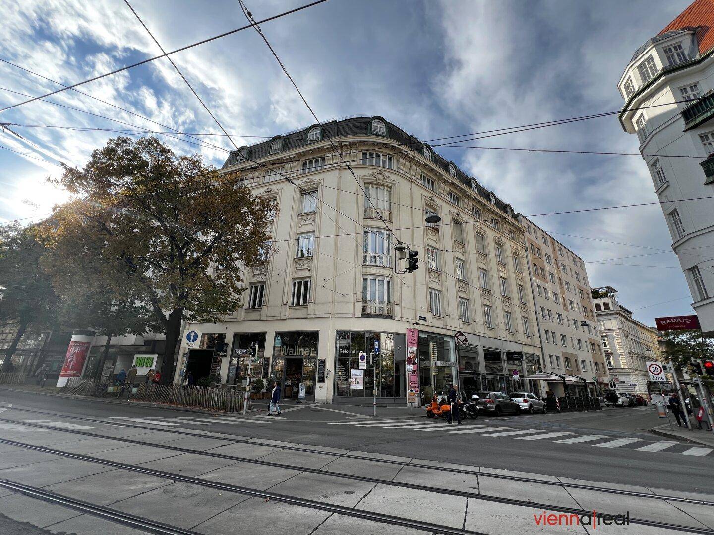 Großes Eckgebäude in der Stadt mit Geschäften und Wohnungen unter blauem Himmel.
