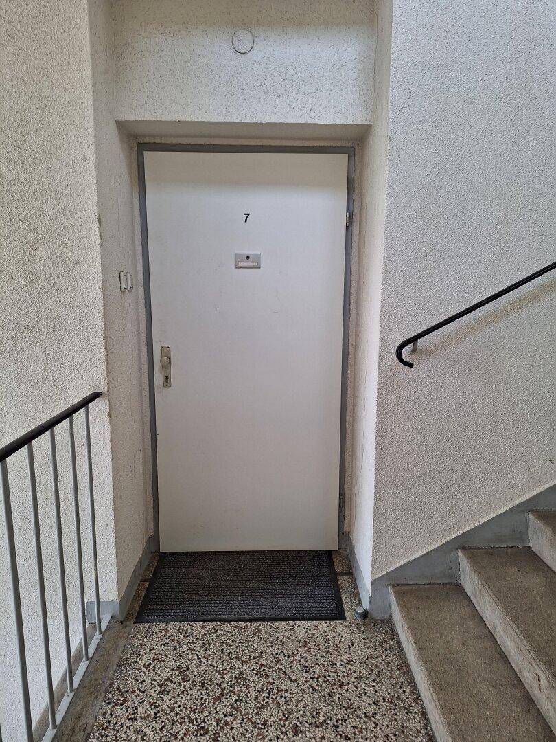 Eingangsbereich mit einer Wohnungstür, die die Nummer 7 trägt, einer Fußmatte und einem robusten Steinboden.