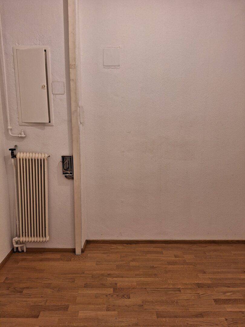 Kleiner Raum mit Heizkörper, Sicherungskasten und einfacher Ausstattung, möglicherweise ein Abstellraum oder Technikbereich.