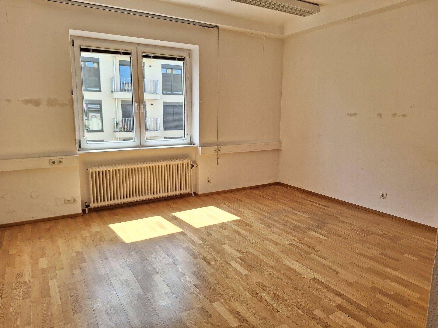 Geräumiger Büroraum mit hellem Parkettboden und einem großen Fenster, das viel natürliches Licht spendet.