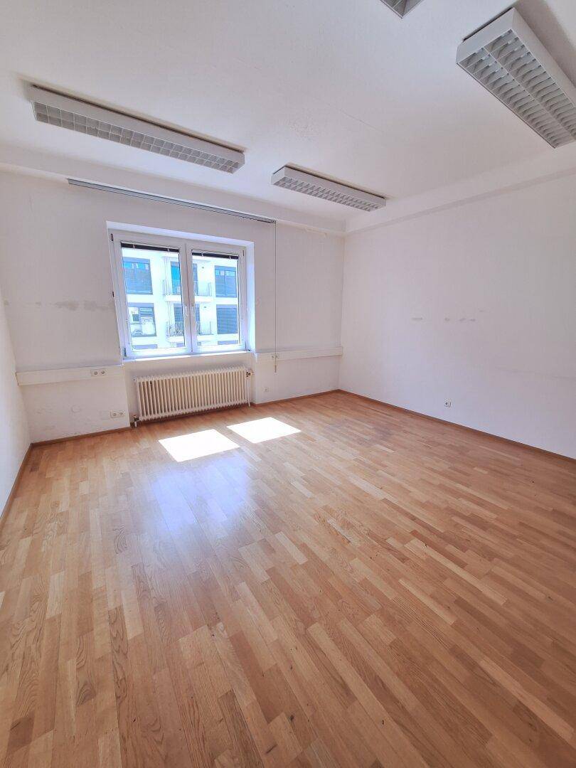 Heller Büroraum mit Parkettboden und einem großen Fenster, das den Raum mit Sonnenlicht durchflutet.