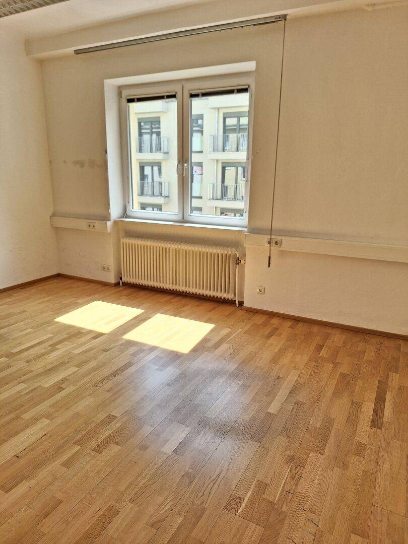 Büroraum mit hellem Parkettboden und einem großen Fenster, das den Raum mit natürlichem Licht erfüllt.